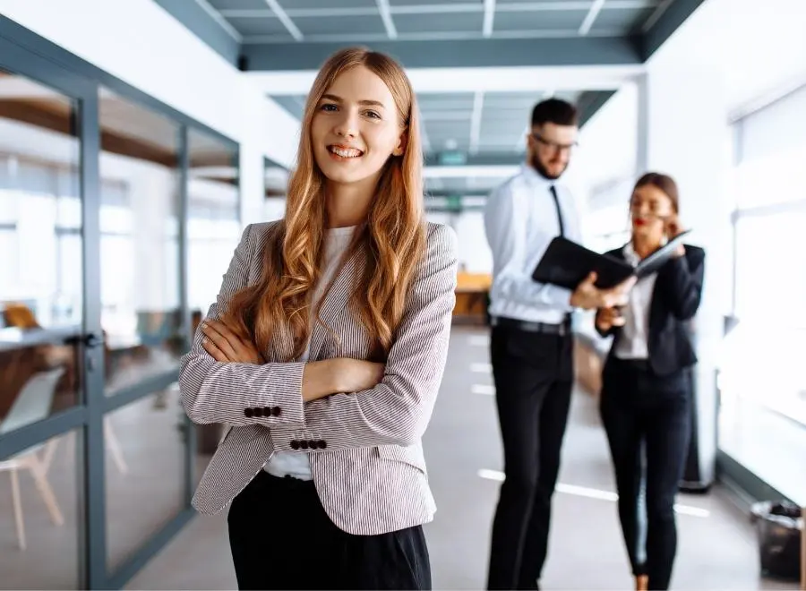 Eine junge Frau im Büro lacht mit verschränkten Armen selbstbewusst in die Kamera.