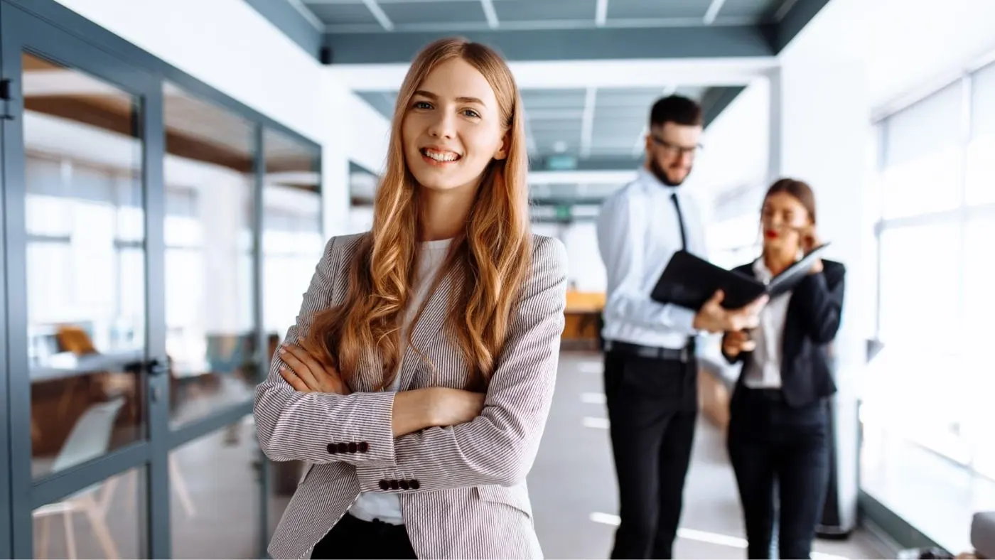 Eine junge Frau im Büro lacht mit verschränkten Armen selbstbewusst in die Kamera.